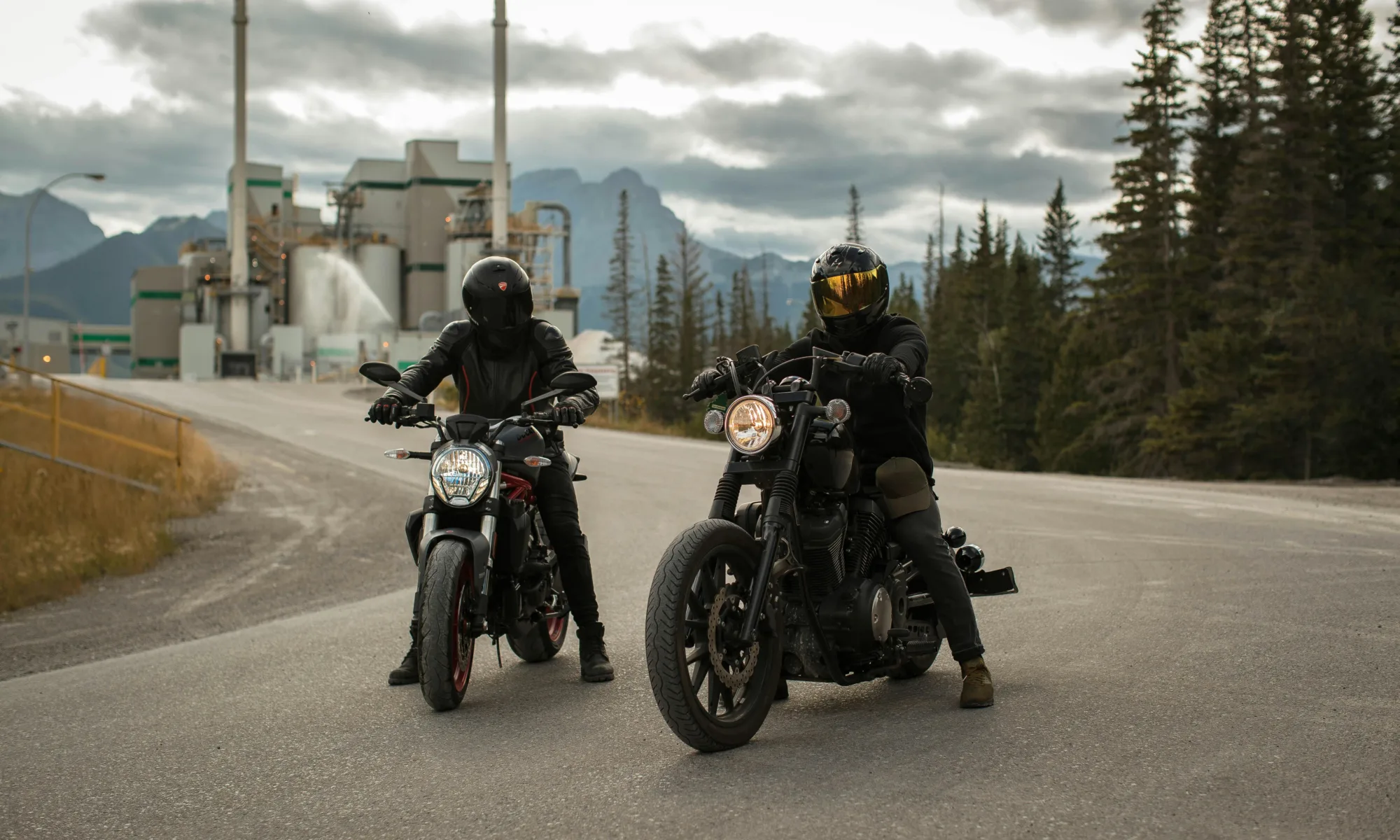 Two motorcyclists riding on a scenic highway with full protective gear, against a backdrop of mountains and pine trees.